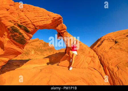 Glückliche Frau entspannende oben in der Arch Rock eine ikonische Natural Arch. Junge kaukasier Rucksack Mädchen im Tal des Feuers, Nevada's älteste State Park berühmt für rote Sandsteinformationen. Sonnenuntergang rote Farbe. Stockfoto