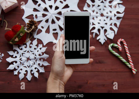 Das Konzept des Neuen Jahres. Eine Frau, die ein Foto für ein Blog. Frauen Hände halten ein Smartphone, Foto Schneeflocken aus Papier, Geschenke, Schere auf einer Stockfoto