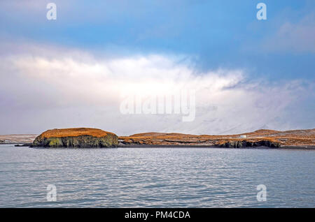 Blick über Reykjavik Bucht zu den grasbewachsenen Hügeln und Felsen der Insel Videy und die Berge dahinter unter einem Himmel mit niedrig hängenden Wolken Stockfoto