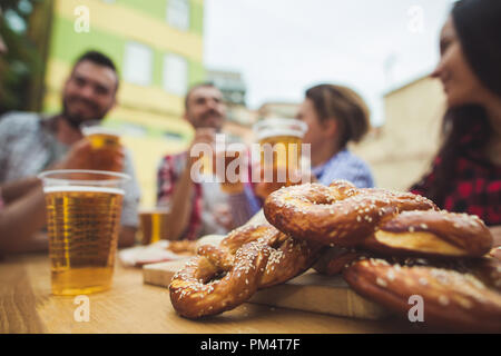 Die Gruppe der Freunde genießen und trinken Bier an der Bar im Freien oder im Pub. Gekocht serviert mit Weißwurst, Bier und Brezeln. Perfekt für Oktoberfest. Natürliche Holz- Hintergrund. Vorderansicht mit weiblichen und männlichen Händen Stockfoto