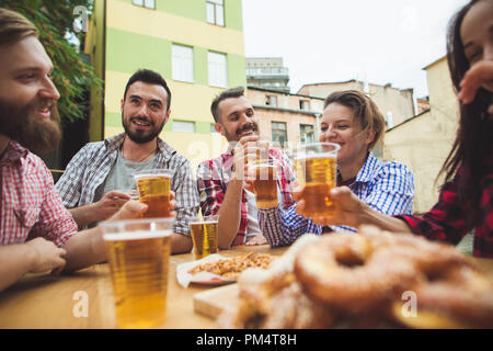 Die Gruppe der Freunde genießen und trinken Bier an der Bar im Freien oder im Pub. Gekocht serviert mit Weißwurst, Bier und Brezeln. Perfekt für Oktoberfest. Natürliche Holz- Hintergrund. Vorderansicht mit weiblichen und männlichen Händen Stockfoto