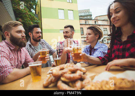 Die Gruppe der Freunde genießen und trinken Bier an der Bar im Freien oder im Pub. Gekocht serviert mit Weißwurst, Bier und Brezeln. Perfekt für Oktoberfest. Natürliche Holz- Hintergrund. Vorderansicht mit weiblichen und männlichen Händen Stockfoto