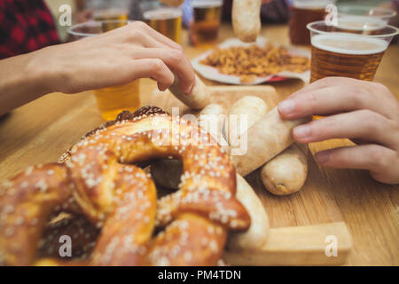 Gekocht serviert mit Weißwurst, Bier und Brezeln. Perfekt für Oktoberfest. Natürliche Holz- Hintergrund. Vorderansicht mit weiblichen und männlichen Händen Stockfoto