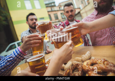 Die Gruppe der Freunde genießen und trinken Bier an der Bar im Freien oder im Pub. Gekocht serviert mit Weißwurst, Bier und Brezeln. Perfekt für Oktoberfest. Natürliche Holz- Hintergrund. Vorderansicht mit weiblichen und männlichen Händen Stockfoto