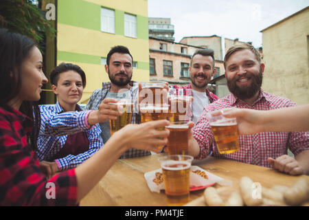 Die Gruppe der Freunde genießen und trinken Bier an der Bar im Freien oder im Pub. Gekocht serviert mit Weißwurst, Bier und Brezeln. Perfekt für Oktoberfest. Natürliche Holz- Hintergrund. Vorderansicht mit weiblichen und männlichen Händen Stockfoto