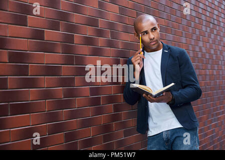 Die Entwicklung des Gehirns. Junger Mann stehend an der Wand lesen Buch berühren Kopf mit Bleistift denken Stockfoto