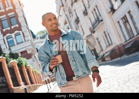 Morgen Energie. Junge Mann zu Fuß auf city street Holding Tasse trinken heißen Kaffee zur Seite schauen, lächeln fröhlicher Stockfoto