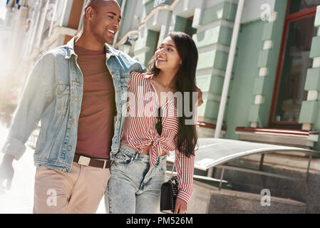 Romantische Beziehung. Junge diverse Paar zu Fuß auf der Straße umarmen sich einander reden Lächeln glücklich Stockfoto