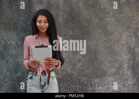 Moderne Technologie. Junge brunette Mädchen stehen auf graue Wand unter Foto auf digital Tablet lächelt glücklich Stockfoto