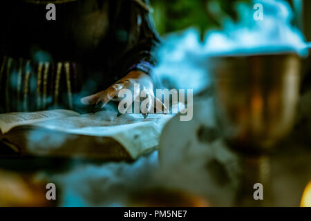 Nahaufnahme der Hand eine Hexe mit Spitzen schwarz Nägel auf smoky Buch der Magie Rezept und mit einem Zauberstab. Stockfoto