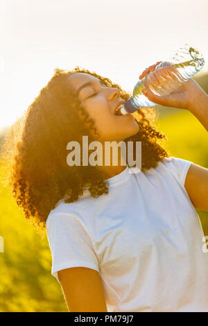 Outdoor Portrait schöne glückliche Mischlinge afroamerikanische Mädchen Teenager weibliche junge Frau Trinkwasser aus einer Flasche in einem Feld von gelben flo Stockfoto