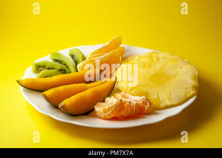 Tropische Früchte, Kiwi, Mango, Ananas, Orange und Mandarine Orange Slices auf weiße Platte auf gelben Hintergrund. Stockfoto