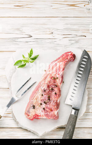 Stück Fleisch Schweinefleisch Brustbein rippen Spachtel mit Salz und Pfeffer gewürzt und Zweig Basilikum auf weissem Holztisch Marmor Stein mit Gabel Messer foo serviert. Stockfoto