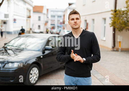 Schöne stilvolle Geschäftsmann in einem schwarzen T-Shirt und blauen Hosen in der Nähe von einem schwarzen Auto Stockfoto