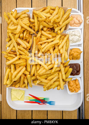 Fünf Portionen Belgische Pommes Frites mit fünf verschiedenen Saucen, Plastikgabeln und Salz Pakete auf einem weißen Kunststoff Fach auf einer hölzernen Tisch. Stockfoto