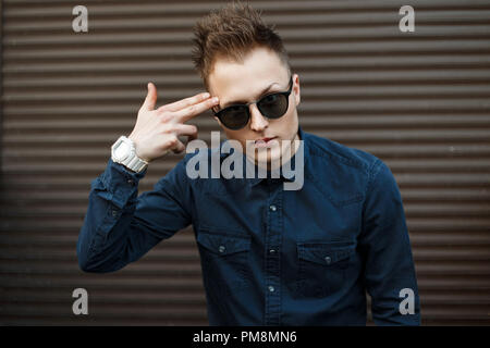 Hübscher junger Mann in Sonnenbrille und ein blaues Hemd zeigt eine Pistole aus seiner Finger in die Nähe von Metall Wand Stockfoto