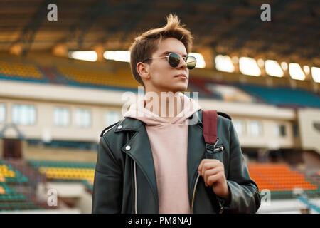 Schöne portrait einer jungen Sport Mann mit Sonnenbrille in einer schwarzen Jacke mit einem Rucksack im Stadion Stockfoto