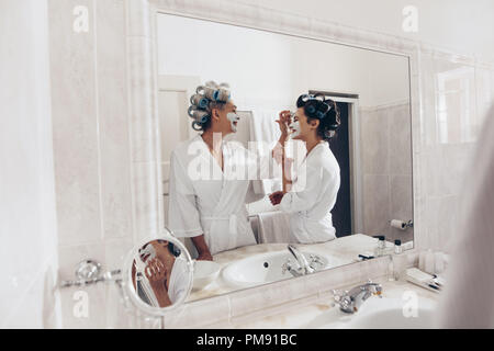 Frauen in Bademänteln und mit Curly Walzen auf Haar Pflege zu Hause. Ältere Frau Anwendung Creme auf das Gesicht ihrer Tochter stand vor der mi Stockfoto