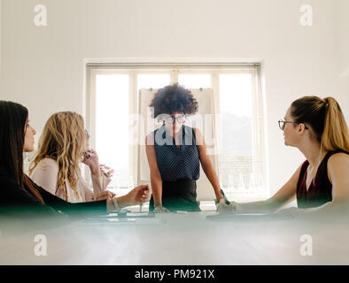 Junge afrikanische Geschäftsfrau, die ihre Ideen teilen zu weiblichen Kollegen sitzen um Konferenztisch im modernen Büro. Gruppe von Frauen in einer Sitzung i Stockfoto
