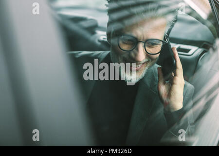 Lächelnd reife Geschäftsmann auf dem Handy beim Sitzen auf der Rückseite der Kabine. Älterer männlichen Pendler mit einem Taxi fahren und das c Stockfoto