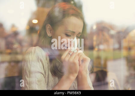 Frau mit kalte Symptome ihre Nase Niesen sitzen in einem Café an einem regnerischen Herbsttag blasen Stockfoto