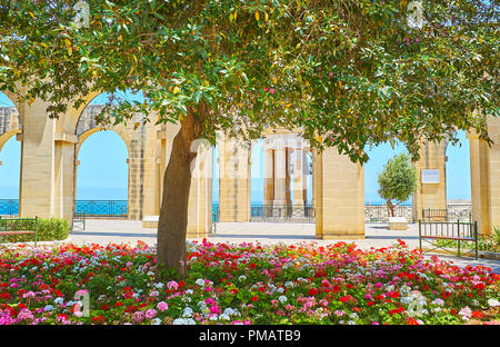 Die malerische gewölbten Wand- und Belagerung War Memorial hinter den üppigen Blumenbeeten und schattenspendenden Baum des Unteren Barrakka Gärten in Valletta, Malta. Stockfoto