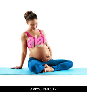 Lächelnd schwangere Frau in Sportkleidung sitzen auf Matten und berühren den Bauch auf weißem Hintergrund. Konzept des gesunden Lebens Stockfoto