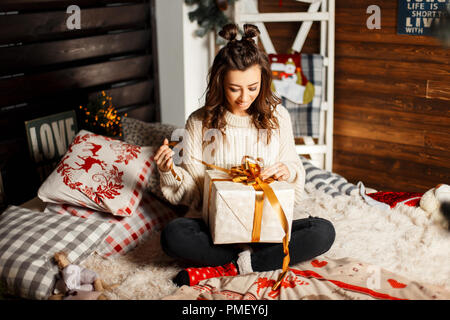 Happy fröhlicher junger Mädchen in einem gestrickten Pullover Vintage öffnet ein Geschenk auf einem Bett am Heiligabend Stockfoto