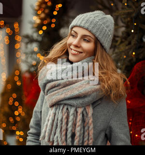 Schöne Frau mit einem Lächeln in einem grauen Mantel Modische Strickmütze und Schal in der Nähe von einem Weihnachtsbaum und Lichter in der Stadt Stockfoto