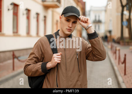 Junge schöne modische Mann in einem stilvollen Vintage Rock mit einer schwarzen Tasche passt seine Kappe auf der Straße Stockfoto