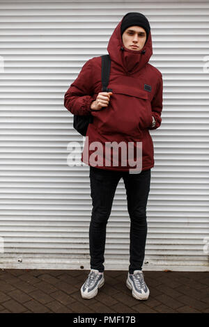 Modische stilvolle Mann in sportlichem Rot Jacke mit einem schwarzen Hut in Weiß modischer sneaker mit einem Rucksack in der Nähe von White Metal Gates auf der Straße Stockfoto