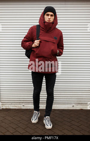 Hübscher junger Mann in Mode Winter Sport Kleidung mit einem Hut und Turnschuhe mit einem Rucksack in der Nähe von white Rollos auf der Straße Stockfoto