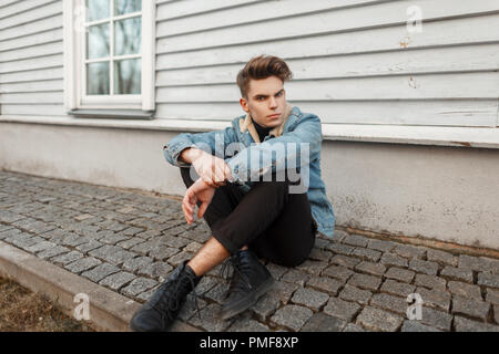 Junge Mode Modell Mann in stilvollen Jeans und schwarze Turnschuhe sitzt in der Nähe von eine Holzwand Stockfoto