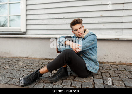 Schöne stilvolle junger Mann mit einem Haarschnitt in einem modischen Vintage Jeans Jacke mit schwarzen Sneakers sitzt in der Nähe von eine Holzwand Stockfoto