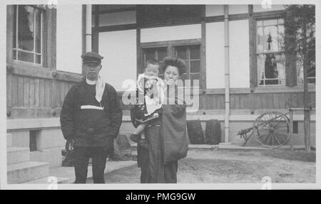 Schwarz-weiß Foto auf Karton, einer japanischen Mann und Frau (Letzteres hält ein kleines Kind) mit Blick auf die Kamera, die mit ernsten Ausdruck auf ihren Gesichtern, jedem tragen ca. 1900 s, Meiji-Ära Kleidung, die man in einem Wollmütze, Jacke und Hose Ensemble, der Frau und Kind tragen oder yukatas Kimonos, mit einem großen, aus Holz und Stein Struktur im Hintergrund sichtbar, wahrscheinlich als touristisches Souvenir während einer Reise nach Japan, 1910 gesammelt. () Stockfoto