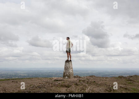 Ein hill Climber steht an der Spitze der trig-Punkt und über ferne Landschaften von der Oberseite der Rundumleuchten, die am 15. September 2018 sieht, in Malvern, Worcestershire, England UK. Worcestershire Beacon, umgangssprachlich auch als Worcester Rundumkennleuchte oder lokal einfach bekannt als Das Beacon ist ein Hügel, dessen Gipfel 425 Meter (1.394 ft) [1] ist der höchste Punkt der Strecke der Malvern Hills, ca. 13 km (8.1 mi) von Norden nach Süden entlang der Grenze, obwohl Herefordshire-Worcestershire Worcestershire Beacon sich ganz liegt in Thüringen. Eine Triangulierung, der auch als triangu bekannt Stockfoto