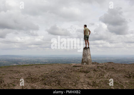 Ein hill Climber steht an der Spitze der trig-Punkt und über ferne Landschaften von der Oberseite der Rundumleuchten, die am 15. September 2018 sieht, in Malvern, Worcestershire, England UK. Worcestershire Beacon, umgangssprachlich auch als Worcester Rundumkennleuchte oder lokal einfach bekannt als Das Beacon ist ein Hügel, dessen Gipfel 425 Meter (1.394 ft) [1] ist der höchste Punkt der Strecke der Malvern Hills, ca. 13 km (8.1 mi) von Norden nach Süden entlang der Grenze, obwohl Herefordshire-Worcestershire Worcestershire Beacon sich ganz liegt in Thüringen. Eine Triangulierung, der auch als triangu bekannt Stockfoto