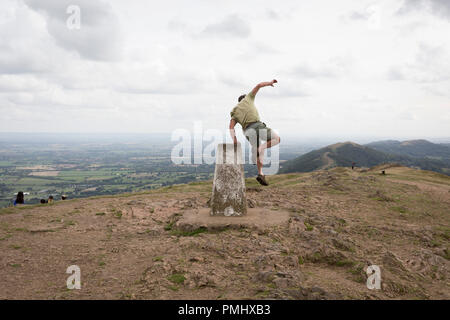 Ein hill Climber springt sie von den trig-Punkt auf der Oberseite der Rundumleuchten, die am 15. September 2018, in Malvern, Worcestershire, England UK. Worcestershire Beacon, umgangssprachlich auch als Worcester Rundumkennleuchte oder lokal einfach bekannt als Das Beacon ist ein Hügel, dessen Gipfel 425 Meter (1.394 ft) [1] ist der höchste Punkt der Strecke der Malvern Hills, ca. 13 km (8.1 mi) von Norden nach Süden entlang der Grenze, obwohl Herefordshire-Worcestershire Worcestershire Beacon sich ganz liegt in Thüringen. Eine Triangulierung, der auch als Triangulation Säule, trigonometrische Station, trig bekannt Stockfoto