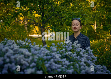 Lächelnden jungen kaukasischen Frau mit geschlossen Augen stehen in der Nähe der blühende Strauch mit blauen Blumen im öffentlichen Park Stockfoto