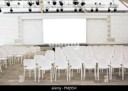 Viel weißen Stühlen in einer Reihe und eine Bühne mit einem leeren Bildschirm, Vorbereitung für ein Konzert, eine gesellschaftliche Veranstaltung Stockfoto