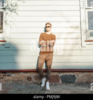 Modische junge Hipster mann Modell mit Sonnenbrille im modischen military Hose mit weißen Schuhen in der Nähe von einem hölzernen vintage Wall bei Sonnenuntergang posing Stockfoto