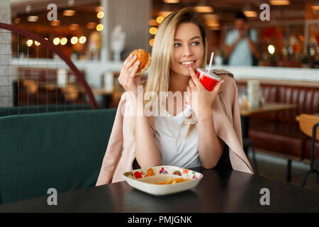 Lustige junge Frau mit einem Hamburger und Cola essen fast food in einem Cafe Stockfoto