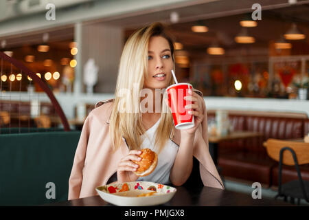 Schöne junge Modell Frau in einem rosa Mode Jacke essen fast food in einem Café, Getränke eine Cola aus einem roten Schale Stockfoto