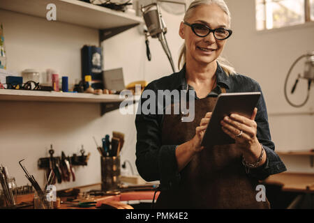 Portrait von Happy reife Frau Schmuck Teekocher mit digitalen Tablet in Ihrer Werkstatt. Lächelnde ältere Frau in goldschmied Workshop mit Tablet-PC. Stockfoto