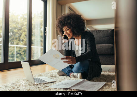 Lächelnde junge Frau sitzt auf dem Boden mit wenigen Berichte Gespräch am Handy. Afrikanische Frauen von zu Hause aus arbeiten. Stockfoto