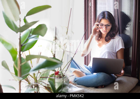 Junge Frau sitzend auf einen Platz am Fenster in der Sonne mit ihren Laptop zu studieren oder Surfen online Stockfoto