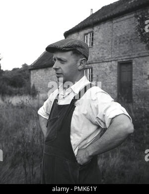 1950, historische, Porträt einer männlichen Immobilien Hausmeister oder Handwerker außerhalb ein Bauernhof, seine Arbeitskleidung, Hemd mit Kragen, dunagarees und flache Kappe tragen, England, UK. Stockfoto