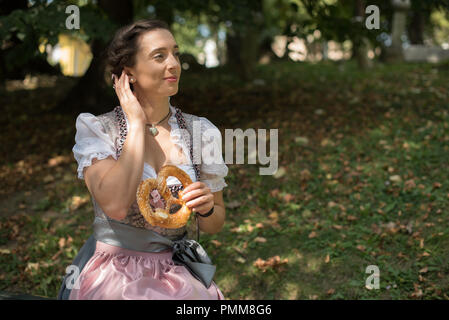 Frau mit einem traditionellen deutschen Dirndl sitzt im Park essen eine Brezel Stockfoto