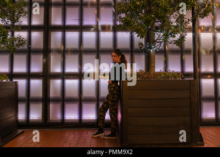 AUCKLAND, Neuseeland - 14. SEPTEMBER 2018 ; Mädchen schiefen Blick nachdenklich auf der Pflanzmaschine vor Zurück panneled beleuchtete Wand in Aucklands Britomart Bereich Stockfoto