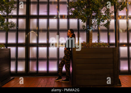 AUCKLAND, Neuseeland - 14. SEPTEMBER 2018 ; Mädchen schiefen Blick nachdenklich auf der Pflanzmaschine vor Zurück panneled beleuchtete Wand in Aucklands Britomart Bereich Stockfoto